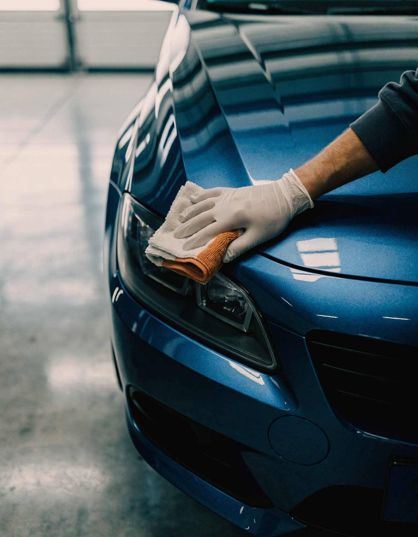 A gloved hand polishing the hood of a blue car in a clean bay.