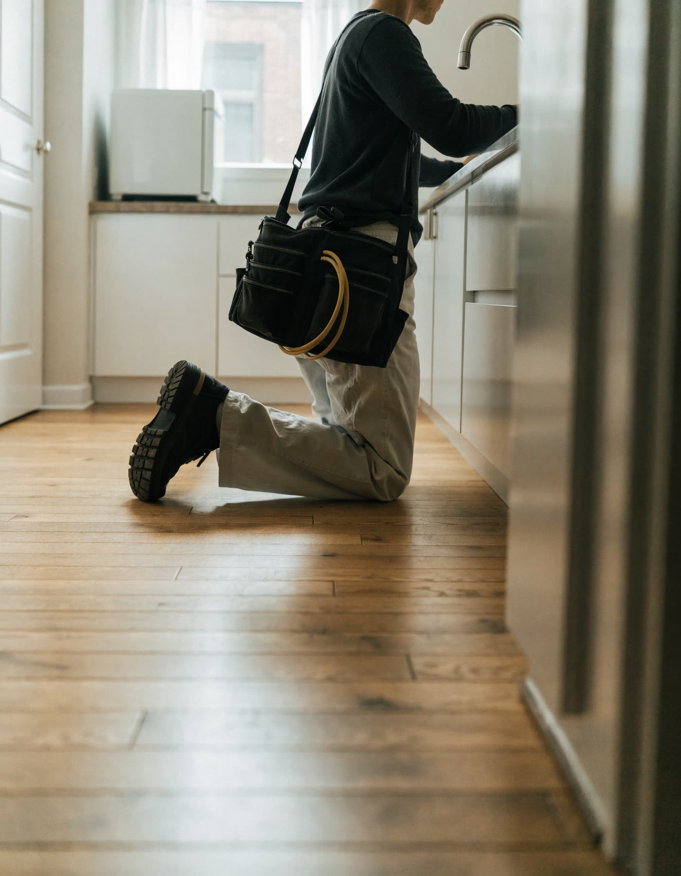 A plumber kneeling at a kitchen sink, tool bag over the shoulder.