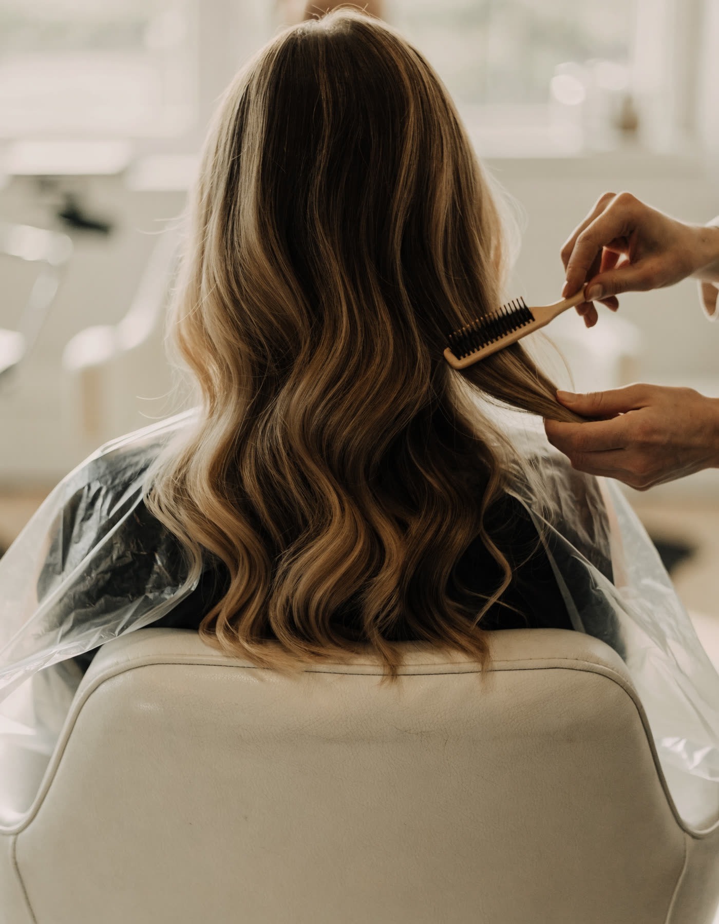 A colourist combing through long balayage hair by a window.